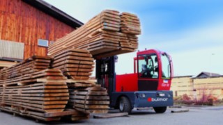 Stapel Holzbretter links vor einer Scheune, rechts der Bulmor EQn 50 Seitenstapler der die geschnittenen Holzbretter aufnimmt