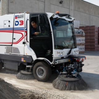 Straßenkehrmaschine Dulevo D6 im Einsatz beim Kehren eines Asphaltbodens, vorbei fahrend an einem Sandhaufen, dahinter eine Lagerhalle aus Beton