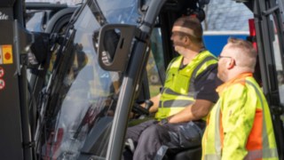 Zwei Mitarbeiter bei der Einweisung im und am Gabelstapler, einer sitzt im Stapler