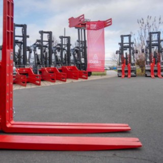 links Gabelzinken in rot auf dem Boden abgelegt, im Hintergrund die Gabelstaplerflotte Linde X-Serie mit Ballenklammer oder verstellbaren Gabelzinken, Farbe der Anbaugeräte ist rot wie die Stapler