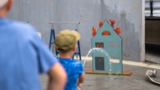 Jugendfeuerwehr Blankenhain Wasserspiel Action Kind was ein Strahlrohr hält und versucht ein brennedes kleines Holzhaus zu löschen